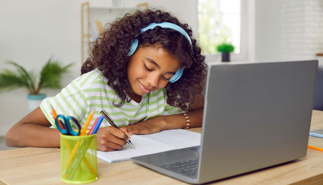 elementary school student in headphones sitting at desk with laptop computer, writing in notebook