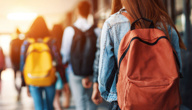 Students with backpacks walk down a sunlit school hallway
