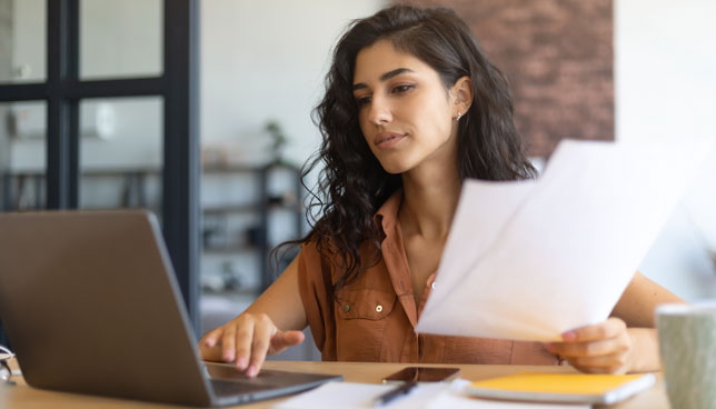 woman working on laptop, holding documents, sitting at desk indoors