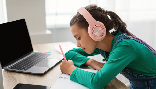 Side view profile of focused little girl writing in a notebook while wearing headphones