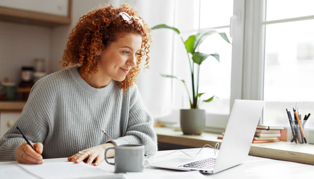 woman with curly red hair sitting at kitchen table with coffee cup and laptop